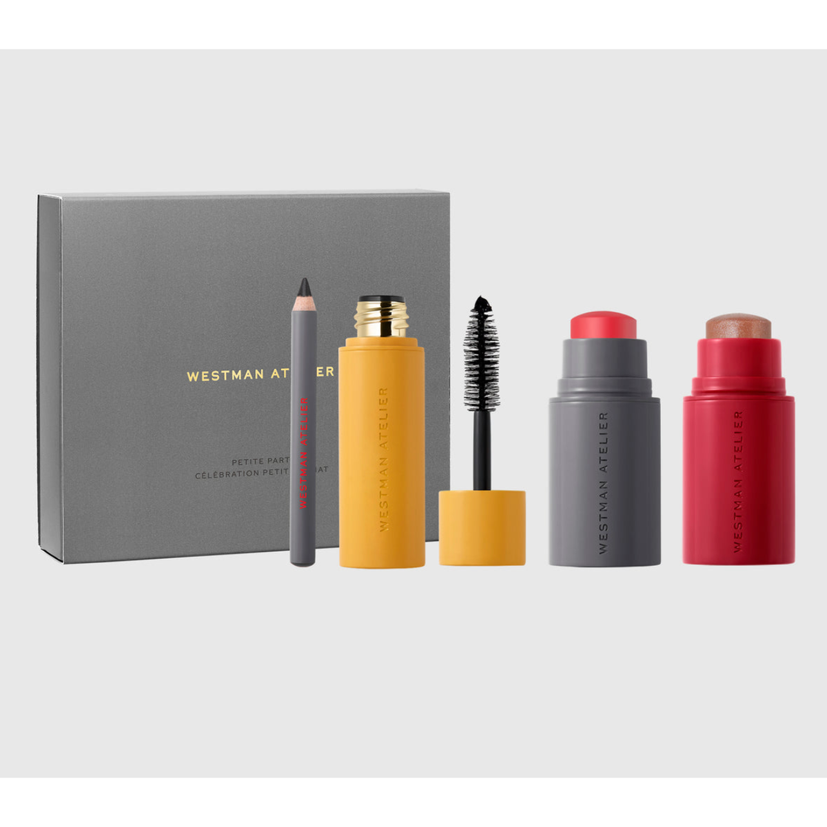 Westman Atelier Petter Faire Celebration Set featuring a gray box, a gray pencil, a yellow mascara tube with wand, a gray lip balm stick, and a red blush stick on a light gray background.