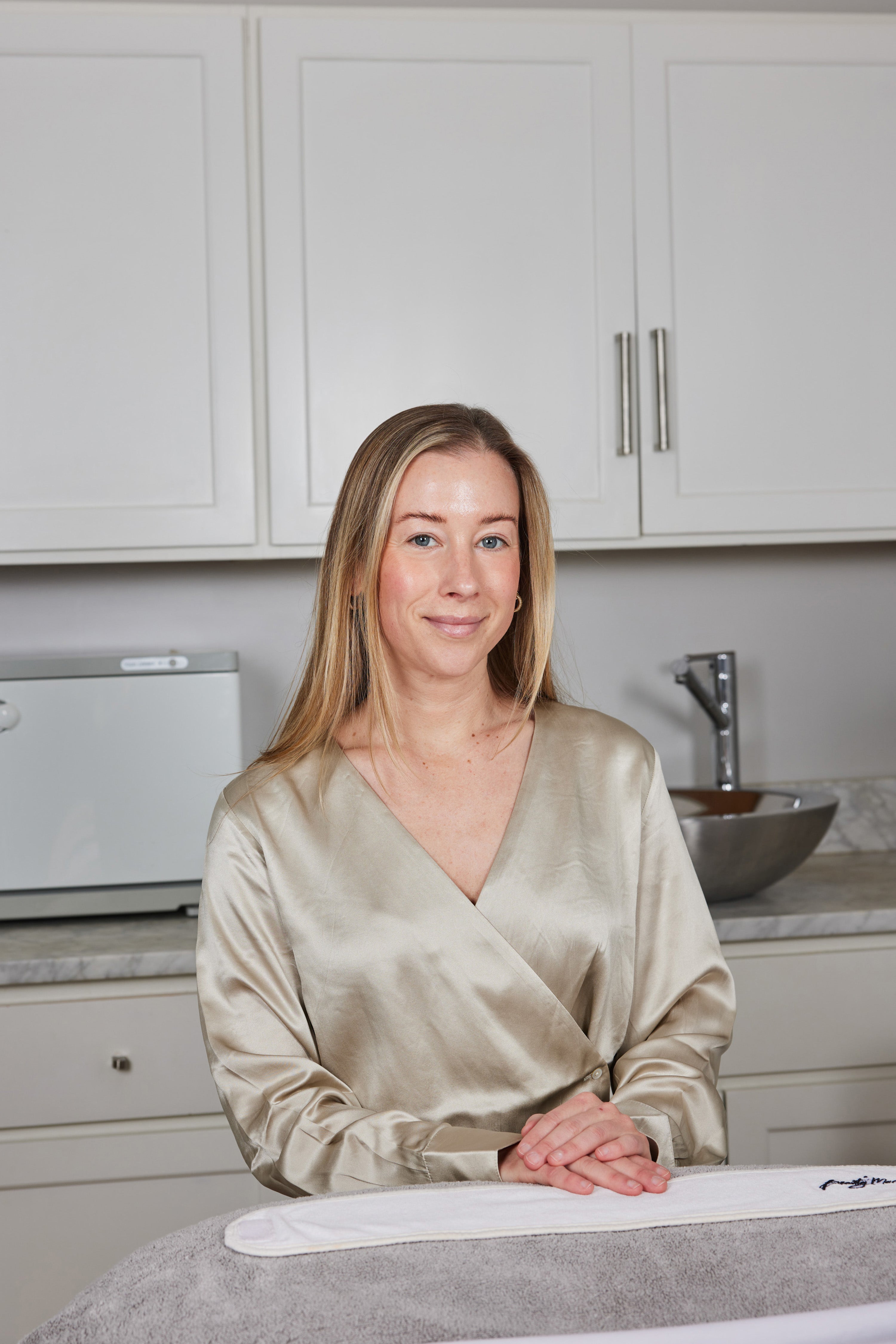 Kristyn, one of our Beauty Mark Estheticians sitting and smiling at the camera wearing a gold satin blouse 
