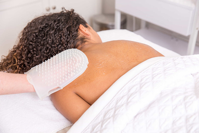 A client lies face down on a white treatment bed with their back exposed while an esthetician, wearing an exfoliating glove, gently massages lotion into the skin