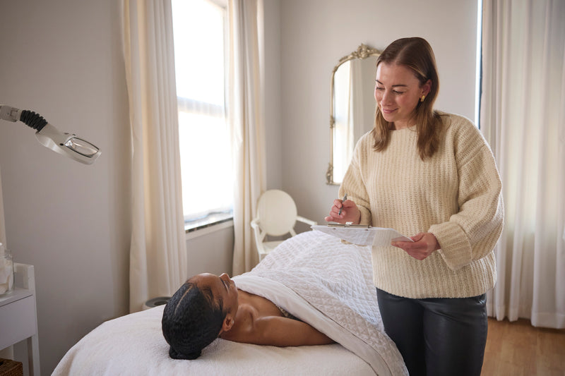 Kristyn, one of our Beauty Mark Esthetician's standing and holding a clipboard while smiling down at client laying in bed for consultation