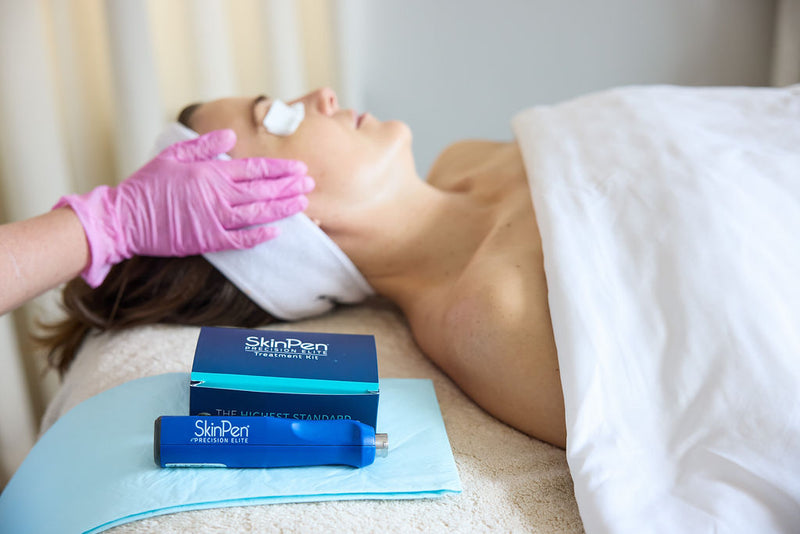 A woman lies on a treatment bed while a gloved hand applies cream to her face. Two blue SkinPen devices and their boxes are placed on a light blue towel in the foreground.