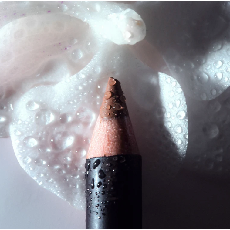 A close-up of a makeup pencil with a pointed brown tip resting on a glossy white surface covered in water droplets.