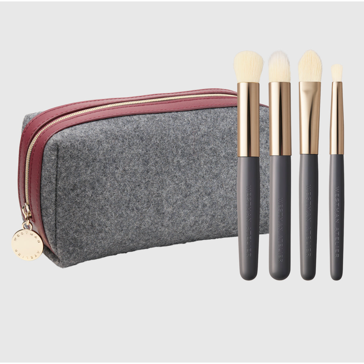 A gray felt makeup bag with a burgundy zipper and gold-tone hardware, shown next to four makeup brushes with dark gray handles, gold ferrules, and cream bristles on a light gray background.