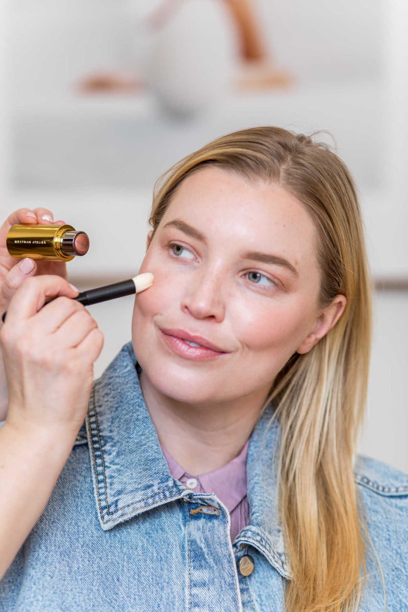 Westman Altelier Lit Up Highlight Stick in Brulee being featured to provide makeup service to a smiling woman with blonde hair